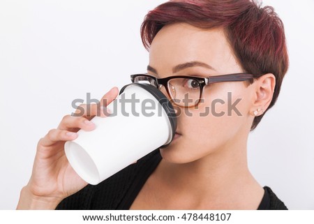 Woman with red hair in wooden glasses with piercing is drinking from coffee cup. Concept of trying to wake up is hard. White background.