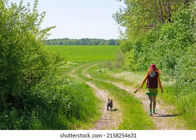 A woman in a red hat walks her French bulldog in nature. The dog is kept on a leash.