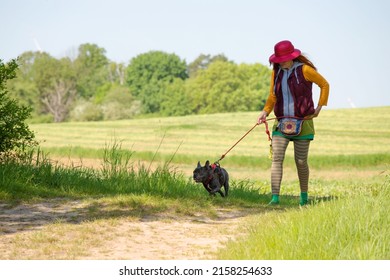 A woman in a red hat walks her French bulldog in nature. The dog is kept on a leash.