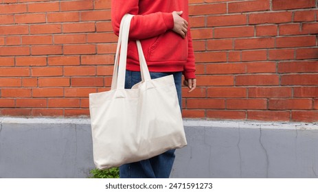 Woman in red hoodie holding tote canvas blank eco bag on street brick wall background. Female consumer hold white textile shopper. template or place for your design, logo, text - Powered by Shutterstock