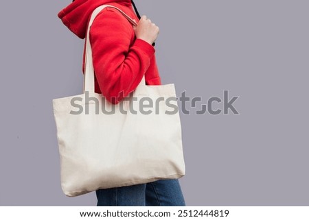 Woman in red hoodie holding tote canvas blank eco bag on street grey minimal wall background. Female consumer hold big white textile shopper. template or place for your design, text, logo