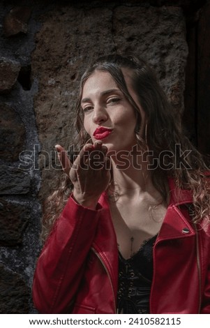 woman in a red jacket, sending a kiss, stands against a textured stone wall, illuminated by natural light