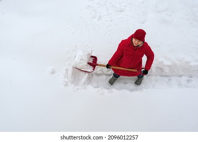 A Woman In A Red Jacket Shovels Her Way Through The Thick Fresh White Snow. Stands With A Snow Shovel And Happily Looks Into The Lens. View From Above. 