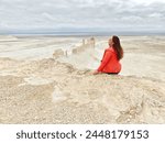 A woman in red jacket sits on a rocky peak overlooking the expansive, arid Bozzhyra landscape. 