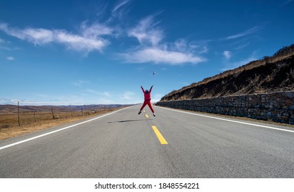 A woman in red jumped into the middle of an empty road.A hat was flying in the air.It's a happy leap.Grass grows on both sides of the road.In the distance are the mountains.