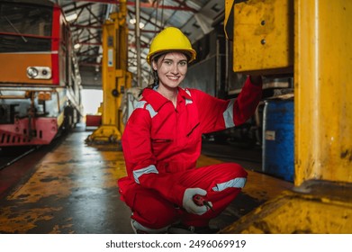 woman in a red jumpsuit is smiling and leaning on a yellow metal pole. She is wearing a yellow helmet and is in a factory setting - Powered by Shutterstock