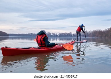 Woman In The Red Kayak And Man Paddle On Stand Up Paddle Boarding (SUP) Rowing On The Danube River On Cloudy Calm Winter Day