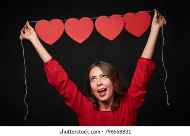 Woman in red lifting above her head a thread with four red heart shapes beaded on it, looking up in excitement, over dark background