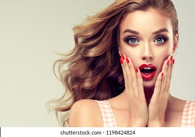 Woman With Red Lips And Nails Surprise Holds Cheeks By Hand .Beautiful Girl  With Curly Hair Surprised And Shocked Looks On You . Presenting Your Product. Expressive Facial Expressions
