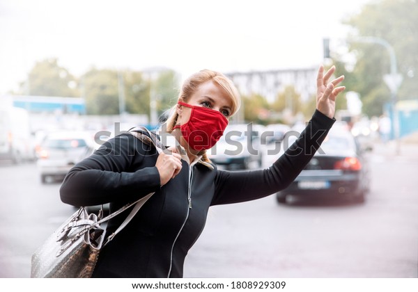Woman with red protective cloth mask on face\
waving to a taxi on City\
street
