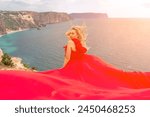 woman red silk dress sits by the ocean with mountains in the background, her dress swaying in the wind.