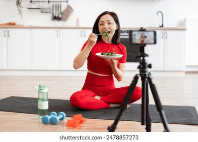 Woman in red sportswear sitting on yoga mat, eating salad while filming fitness vlog in modern kitchen. Healthy lifestyle, nutrition, home workout concepts. - Powered by Shutterstock