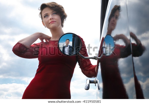 Woman in red stands behind the mirror of the\
car in which reflacts man in blue\
suit