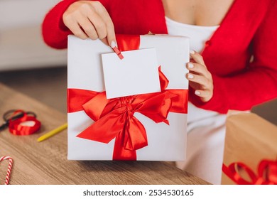 A woman in a red sweater holds a beautifully wrapped gift box with a red ribbon. A blank tag is attached. The scene is set on a wooden table, evoking a festive atmosphere. - Powered by Shutterstock