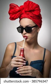 Woman with red turban and sunglasses drinking from a bottle with a straw. Attractive girl portrait holding a bottle, studio shot on gray background. Happy young female, advertisement concept