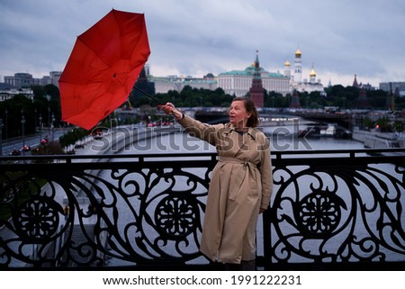 A woman with a red umbrella stands on a bridge against the background of the Kremlin in Moscow, wind and bad weather