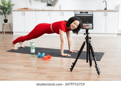 Woman in red workout clothes exercising on mat recording fitness video smiling in modern kitchen setting.Concept of healthy living and home workout. - Powered by Shutterstock