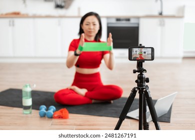 Woman in red workout clothes filming online fitness class at home using resistance band. Fitness instructor demonstrating exercises for virtual audience. Home workout setup with tripod, smartphone. - Powered by Shutterstock