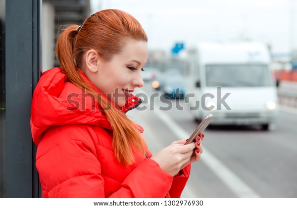Woman, redhead girl in\
side profile holding a phone, texting an sms outside on a bus\
station, near the road in red winter coat clothing. Ordering a taxi\
concept.