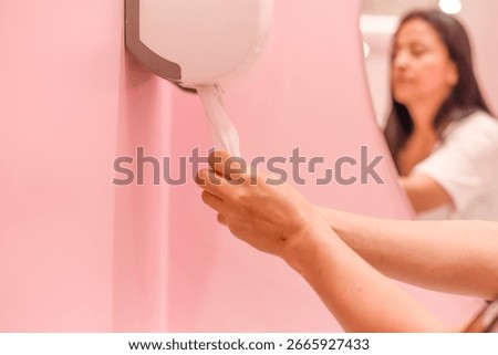 Woman reflected in mirror while pulling paper towel from dispenser in pink restroom, emphasizing hygiene habits