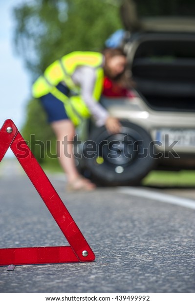 Woman in reflective vest changing tire on\
the roadside. Focus on red emergency\
sign