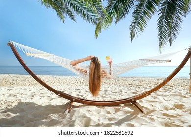 Woman with refreshing cocktail relaxing in hammock under green palm leaves on beach - Shutterstock ID 1822581692