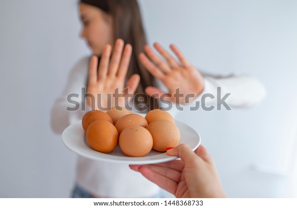 Woman refusing to eat eggs. Egg Free Affected\
Allergy Banned Restriction. Young beautiful woman holding fresh egg\
at home with open hand doing stop sign with serious and confident\
expression