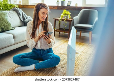 Woman regulating heater temperature. Elegant woman in beige blouse in the modern house in sunny winter day sitting near couch and white electric radiator using smartphone app for temperature control. - Powered by Shutterstock