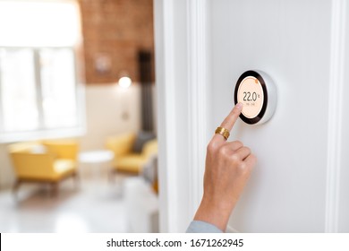 Woman regulating heating temperature with a modern wireless thermostat installed on the white wall at home. Cropped view focused on hand