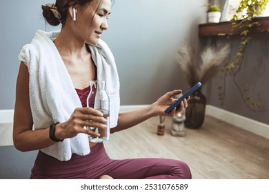 A woman relaxes after her yoga session, holding a phone and a glass of water. She is dressed in workout attire with a towel draped over her shoulders, exuding a sense of calm. - Powered by Shutterstock