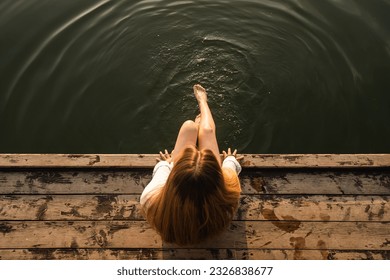 Woman relaxes by the lake sitting on the edge of a wooden jetty, swing feet near the water surface, top view.
