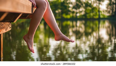 Woman relaxes by the lake sitting on the edge of a wooden jetty , swing one's feet near the water surface. Sunny joyful summer day or evening concept. 