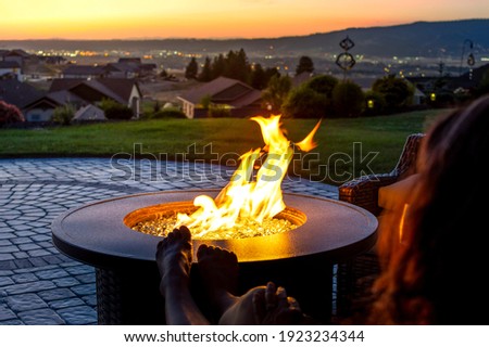 A woman relaxes by a roaring firepit on a paver patio at sunset overlooking the Spokane Valley, in Washington State, USA