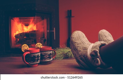 Woman relaxes by warm fire with a cup of hot drink and warming up her feet in woollen socks. Close up on feet. Winter and Christmas holidays concept. Cup with Christmas ornament near fireplace. 