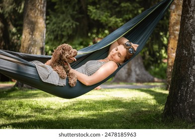 Woman relaxes in hammock with her fluffy brown Maltipoo dog on sunny day. Both looking content and happy. This serene outdoor scene captures joy of bonding with pets and enjoying nature together. - Powered by Shutterstock