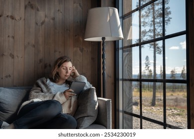 A woman relaxes on a sofa while reading on her E-Reader, in a comfortable wool vest enjoying the natural light streaming through the window in a scenic cabin setting in a forest - Powered by Shutterstock