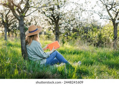 Woman is relaxing in blooming orchard and reading book to improve her mindfulness and mental health. Digital detox outdoors at springtime