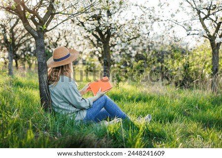 Woman is relaxing in blooming orchard and reading book to improve her mindfulness and mental health. Digital detox outdoors at springtime