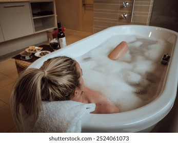 A woman relaxing in a bubble bath with wine and snacks nearby in a modern bathroom - Powered by Shutterstock