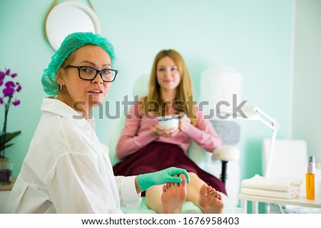 Woman relaxing in chair with cup of coffee, having foot treatment in spa salon.  Professional medical pedicure specialist in protective mask using special electric lathe and tools. 