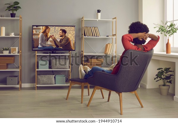 Woman relaxing in chair at home and enjoying\
television soap opera with a happy ending. Young lady sitting in\
comfortable armchair in living-room and watching romantic series or\
reality show on TV