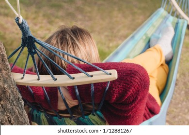 Woman Relaxing In Colorful Striped Textile Hammock Outdoors In Garden. Back View, Warm Clothes, No Shoes