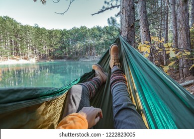 Woman relaxing in the hammock by the lake in the forest, POV view of legs in trekking boots. Hiking in cold season. Wanderlust concept scene.