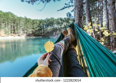 Woman Relaxing In The Hammock By The Lake In The Autumn Forest, POV View Of Legs In Trekking Boots. Hiking In Fall. Wanderlust Concept Scene.