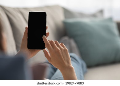 Woman relaxing at home on couch and surfing on internet in free time. Young lady uses smartphone with empty screen, typing message, watch video in living room interior, view over shoulder, cropped