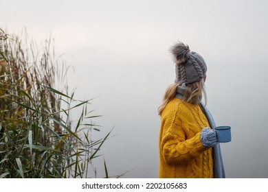 Woman Relaxing With Hot Drink Next To Lake At Autumn Cold Morning. Camping Outdoors At Fall Season. Blond Hair Woman Wearing Knit Hat And Yellow Sweater