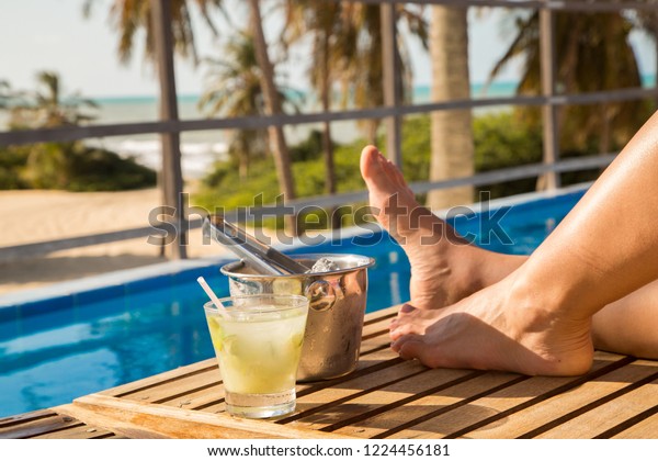 Woman relaxing on a Brazilian beach in sunny day with a caipirinha glass on the side. Only the feet appearing in the photo. In the background coconut palms and a beautiful beach in northeastern Brazil