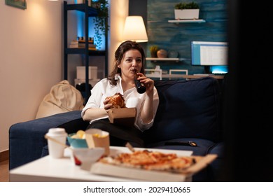 Woman Relaxing On Couch Holding Tasty Delicious Burger While Drinking Beer Watching Entertainment Movie Series On Television. Caucasian Female Enjoying Takeaway Food Home Delivered At Night