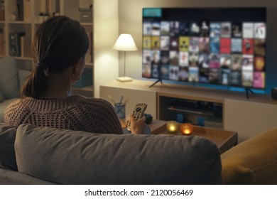 Woman Relaxing On The Couch, She Is Using The Remote Control And Choosing A TV Show Or Movie On The Television Menu