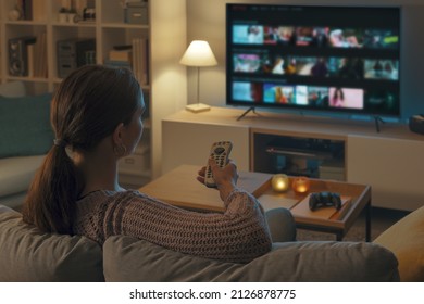 Mujer relajándose en el sofá, está usando el mando a distancia y eligiendo un programa de televisión o una película en el menú de la televisión